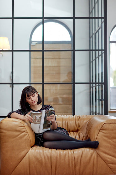 Woman reading a magazine on a brown leather couch in a modern room with large windows wearing black 30 denier opaque tights