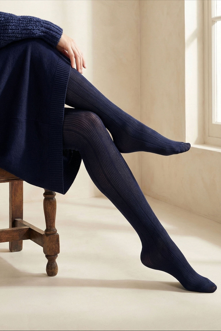 Person sitting on a wooden chair, wearing a navy jumper dress and navy tights