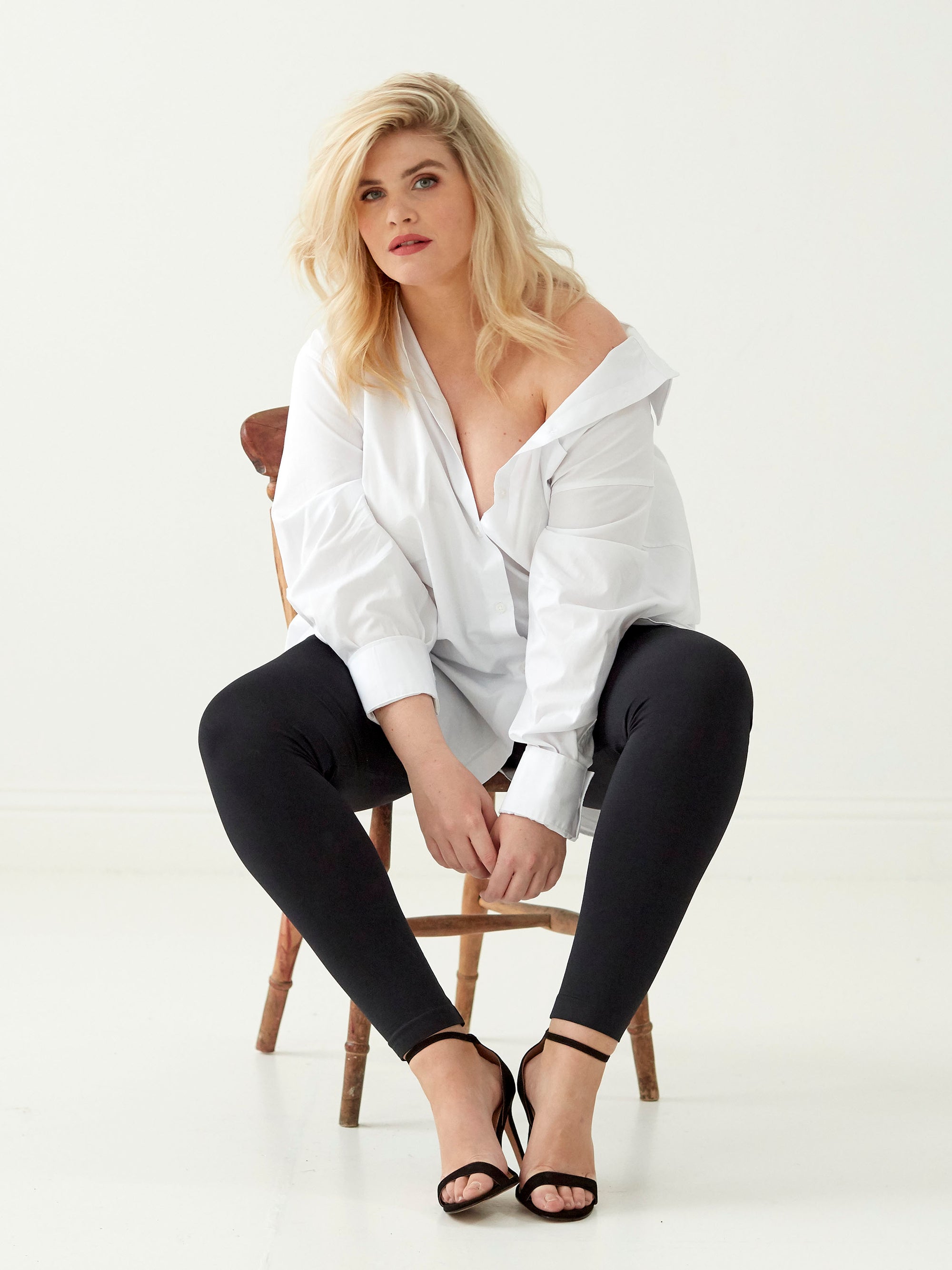Woman wearing a white shirt and black leggings sitting on a chair against a white background