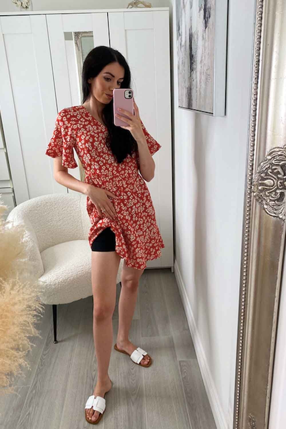 Model taking a mirror selfie wearing a red floral dress and black shapewear shorts in a room with white walls and a chair.