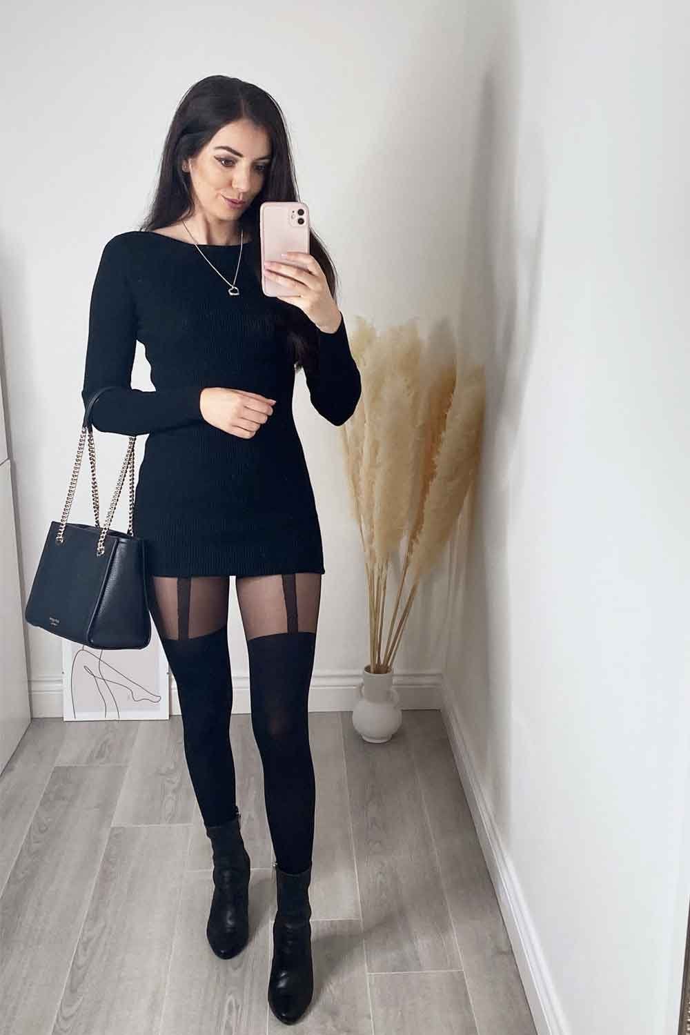 Model taking a mirror selfie wearing a black dress and mock suspender tights  in a room with a white wall and decorative plant.