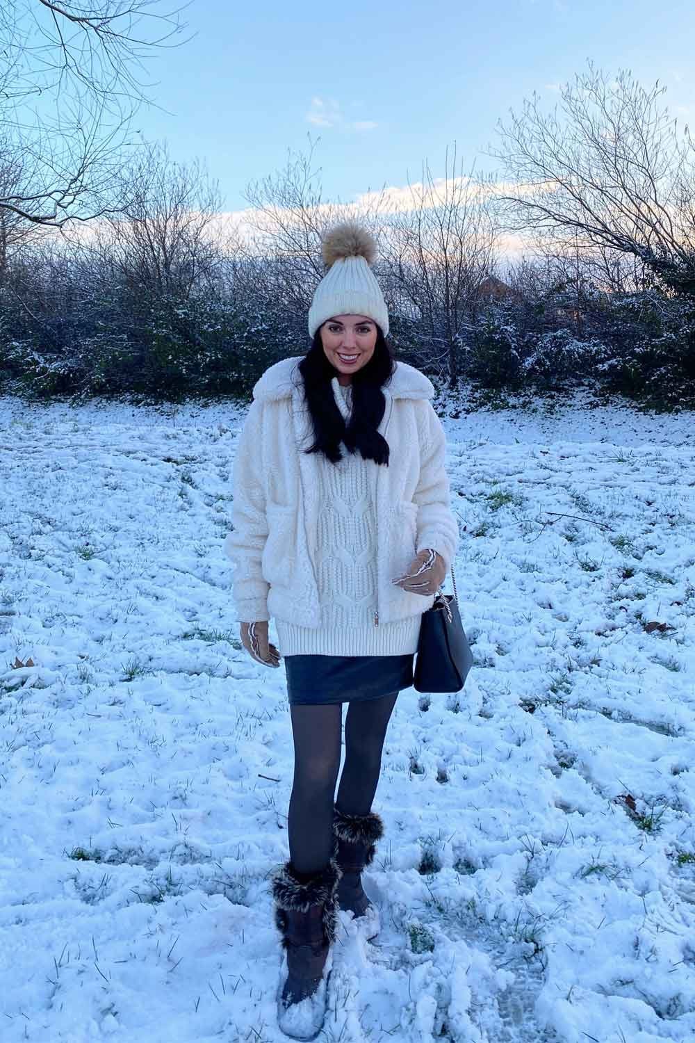 Model wearing a white knit sweater with  sheer fleece lined tights  and hat standing in a snowy field.