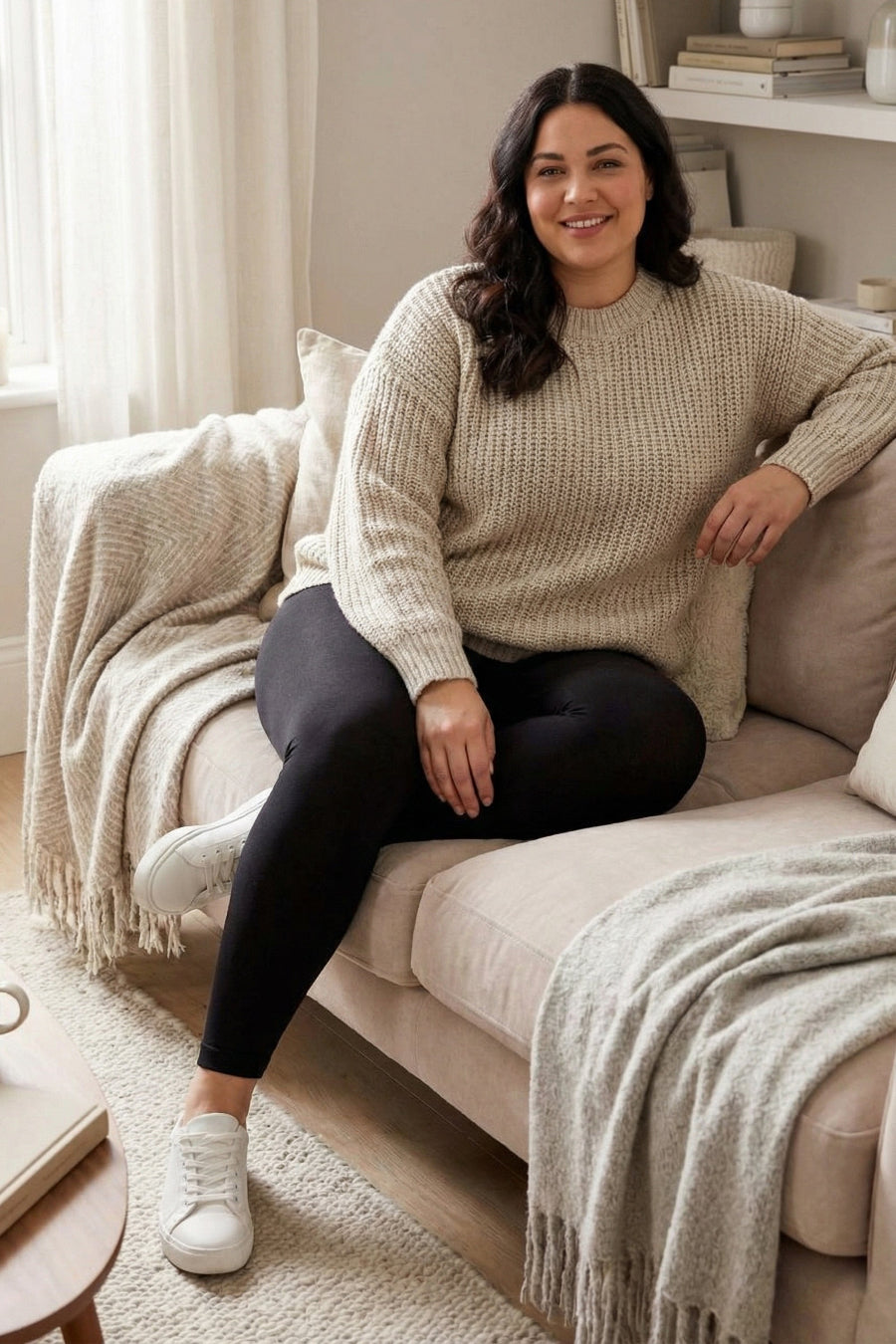 Woman sitting on a couch wearing a beige sweater and black leggings.