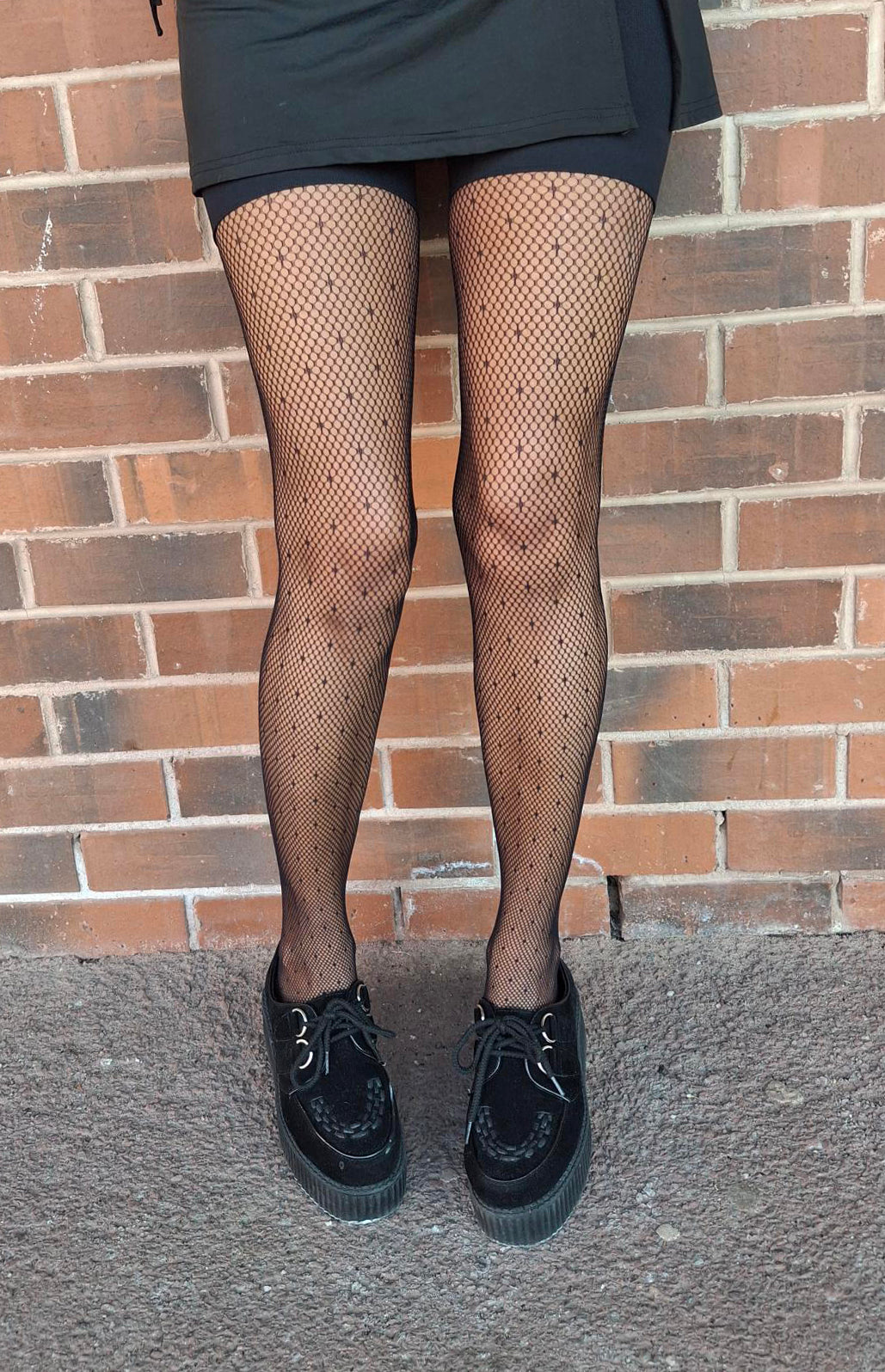 Person wearing fishnet tights and black shoes in front of a brick wall.