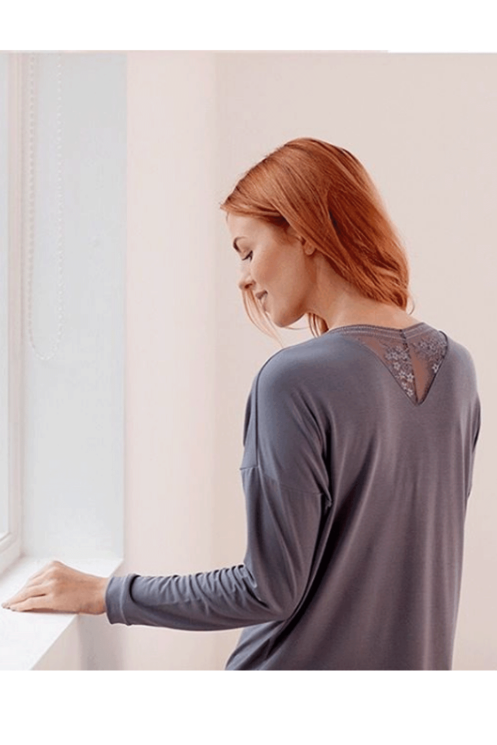Woman wearing a nightshade colour long-sleeve top with lace detailing on the back, standing by a window.