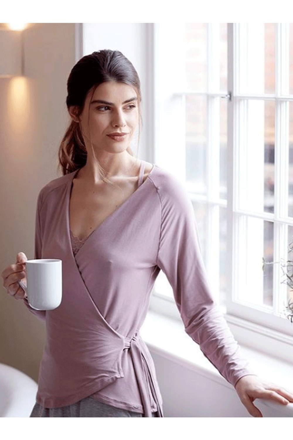 Woman in a pink wrap top  holding a white mug by a window.