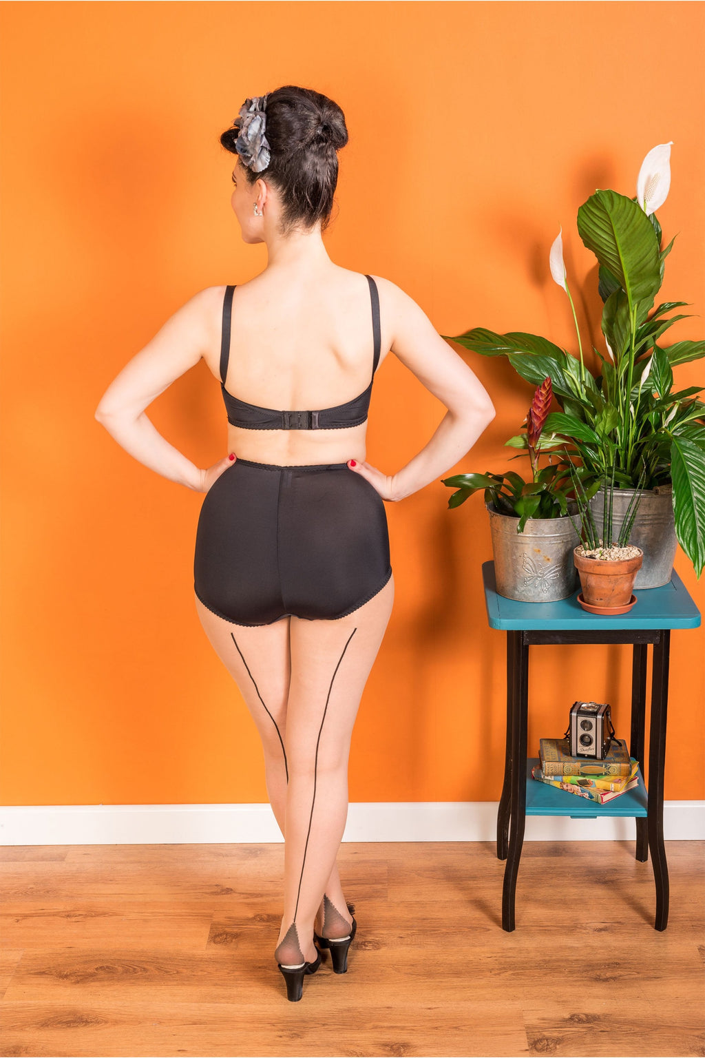 Woman wearing black lingerie and seamed tights standing in front of an orange wall with a small table and plants.