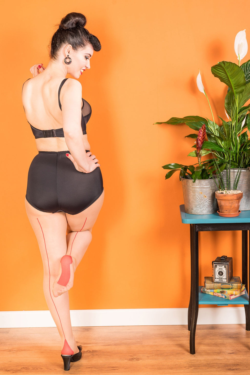 Woman in black lingerie and seamed tights standing against an orange wall with a plant and small table in the background.