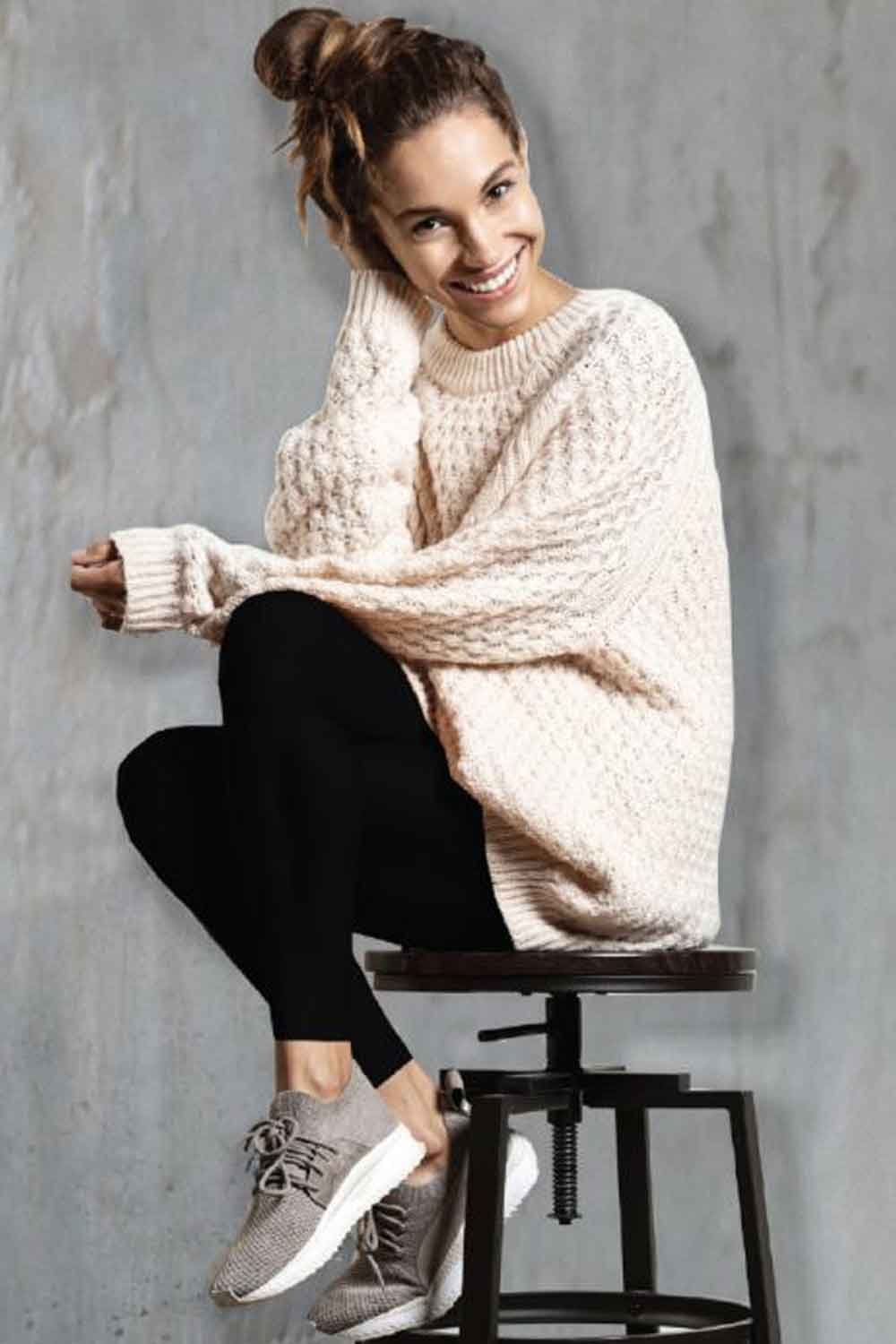 Woman wearing a large beige sweater and flecce lined footless tights sitting on a stool against a grey wall