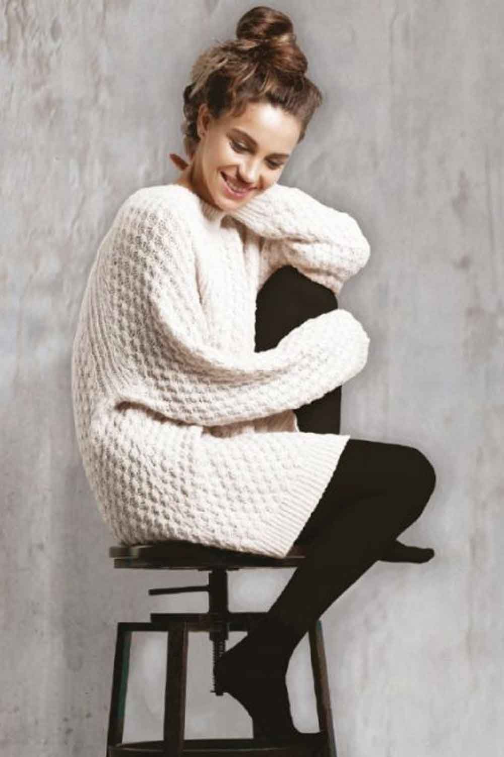Woman wearing a white chunky knit sweater and fleece lined tights  sitting on a stool against a gray wall.