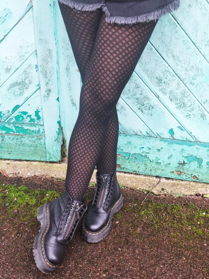 Person wearing black patterned  tights and black boots in front of a green wooden door.