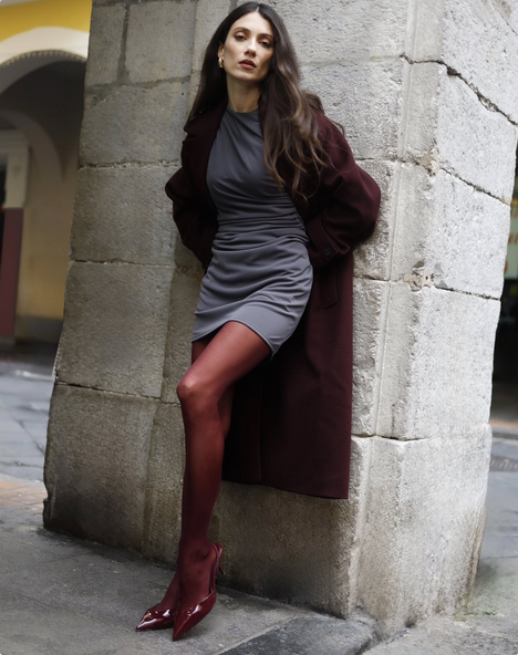 Woman in a gray dress and dark cherry coat and dark cherry tights leaning against a stone wall.