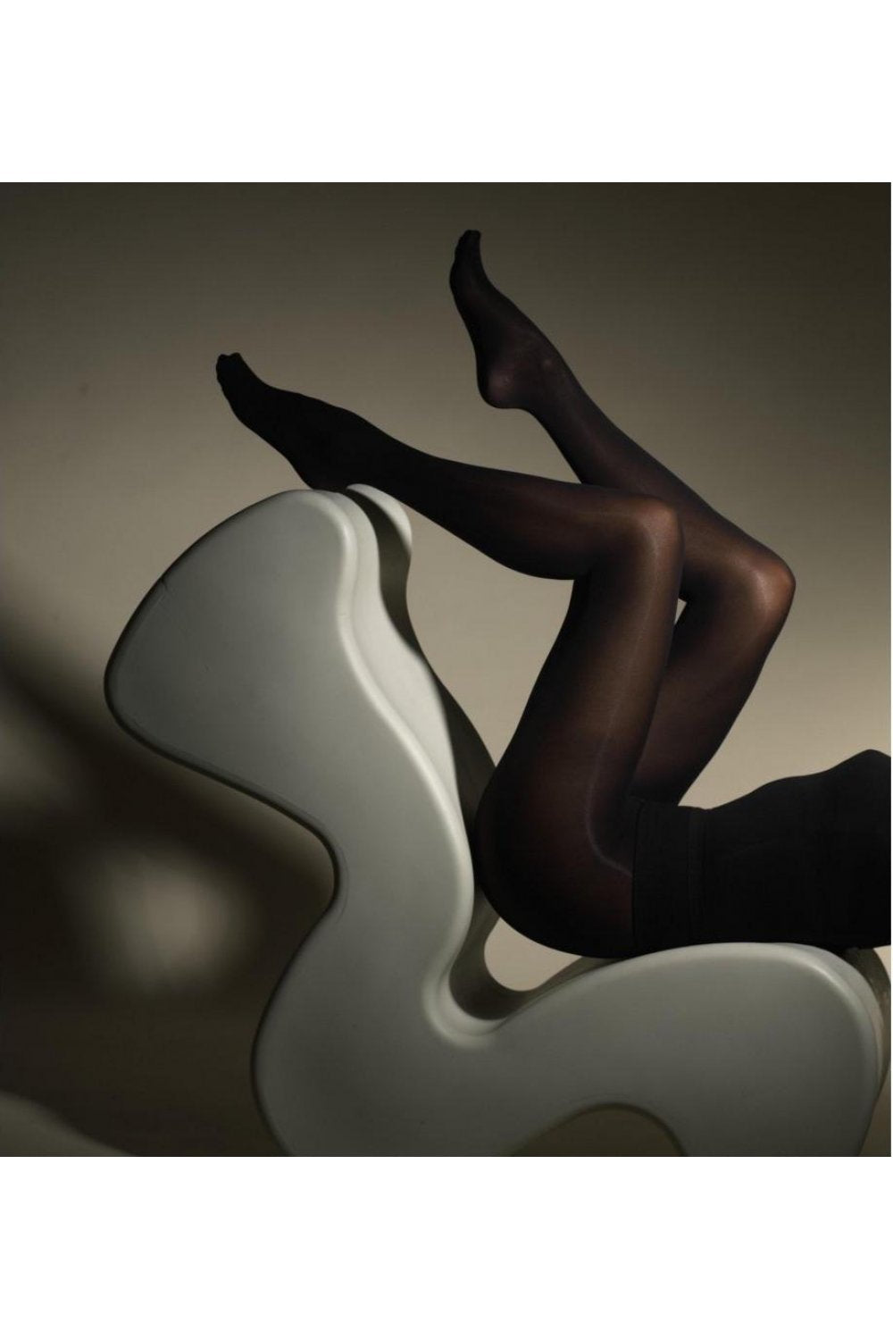 Model wearing black tights sitting on a modern white chair against a neutral background