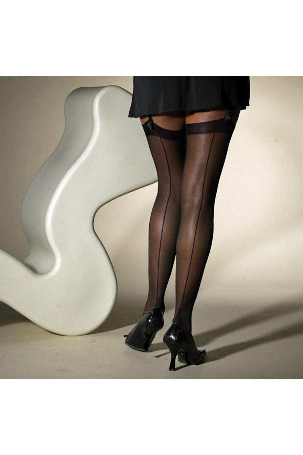 Model wearing black sheer seamed  stockings and high heels in front of a white chair. 