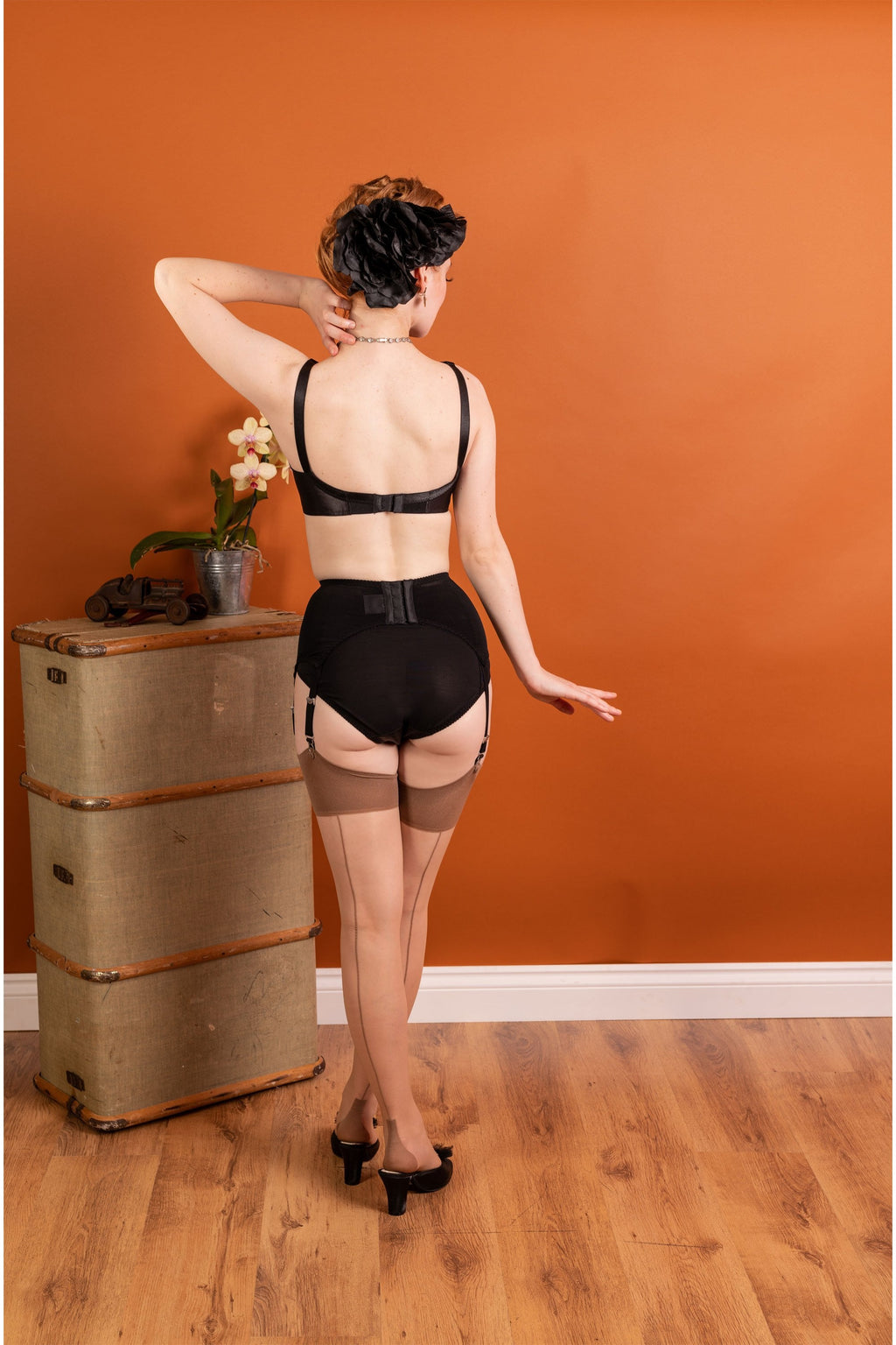 Person wearing a black lingerie and seamed stockings against an orange wall.
