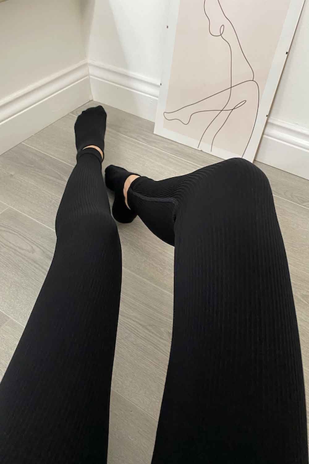 Model wearing black ribbed  leggings and black socks  on a light wooden floor with a white wall and abstract art in the background.
