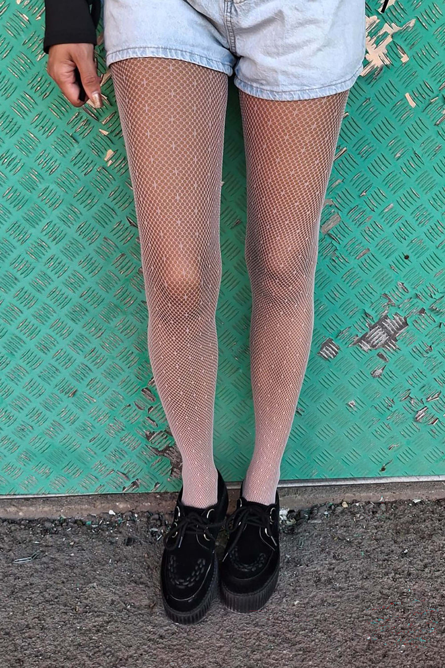 Person wearing fishnet tights and black shoes against a green tiled wall.