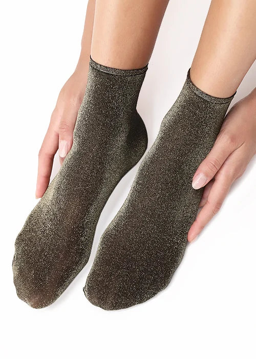 A pair of ankle socks in a shimmering black and gold glitter fabric, shown on a person's feet with hands gently holding the sides against a clean white background.