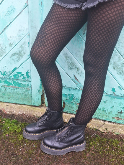 Person wearing black patterned  tights and black boots against a textured blue wall.