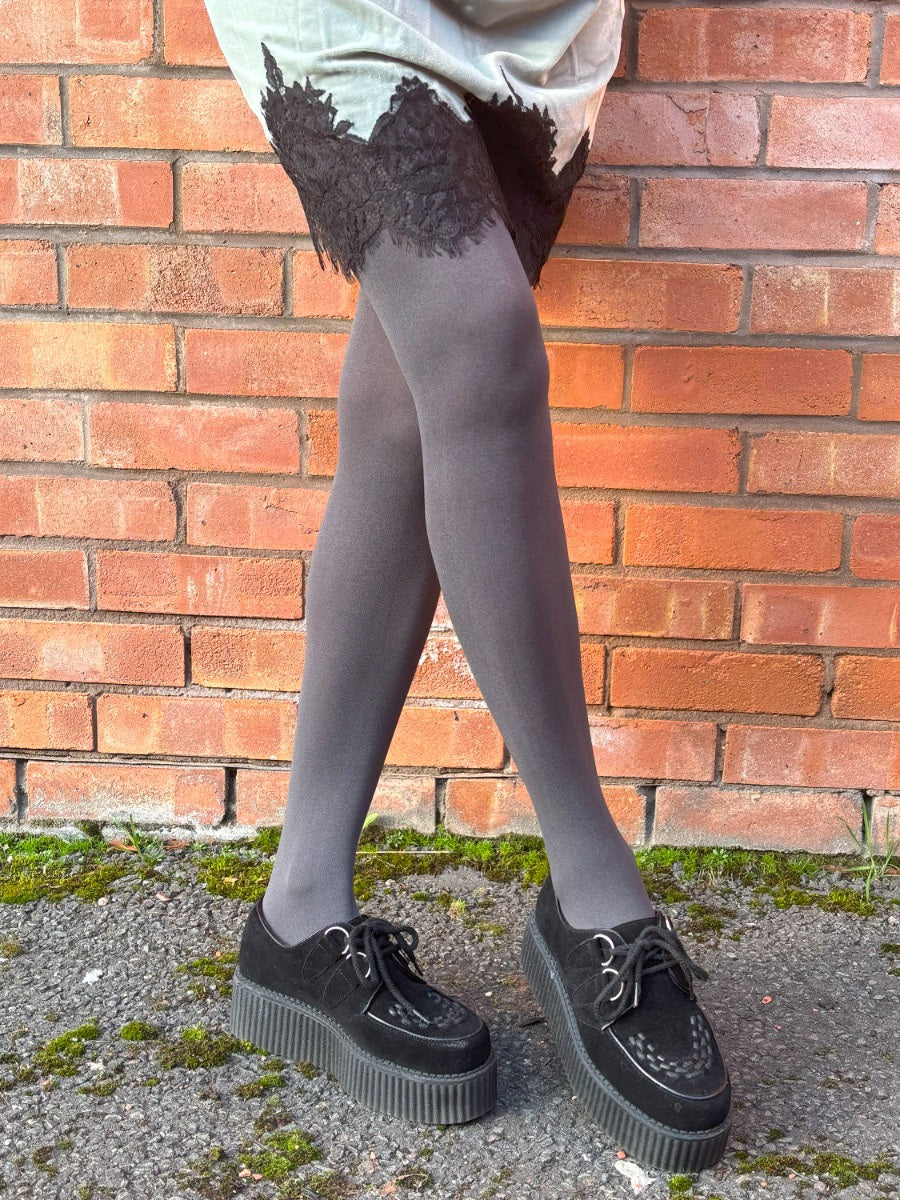 Person wearing black shoes and grey tights standing against a brick wall.