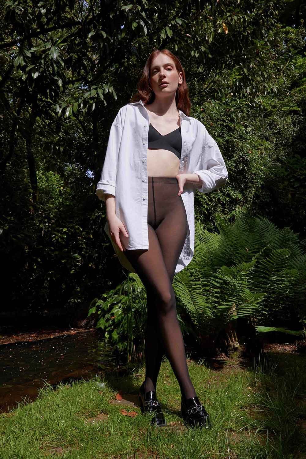 Woman wearing a white shirt, black bra, and black tights standing outdoors with greenery in the background