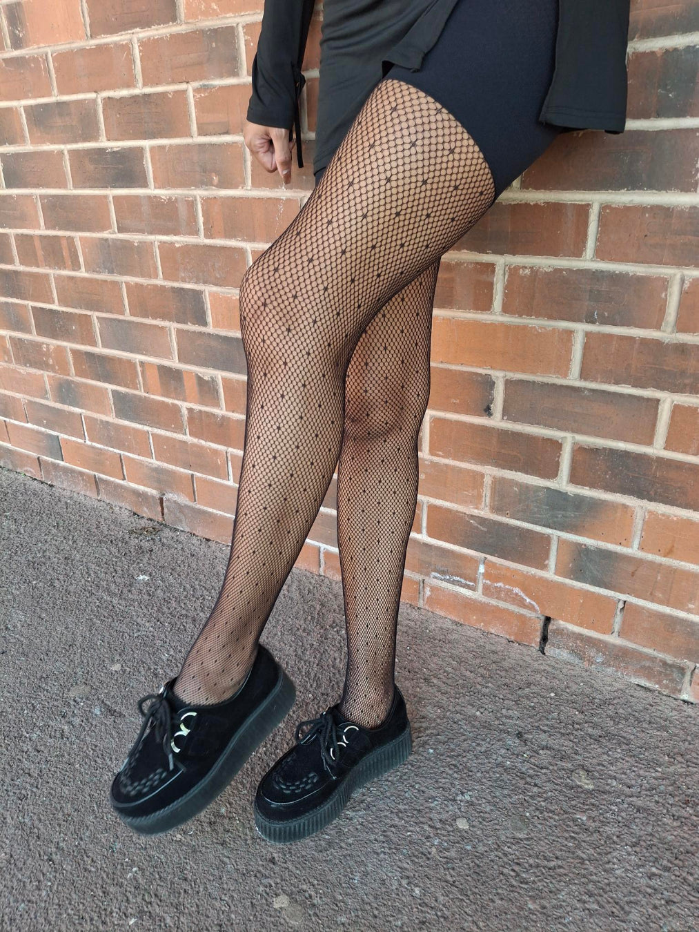Person wearing fishnet tights and black shoes against a brick wall.