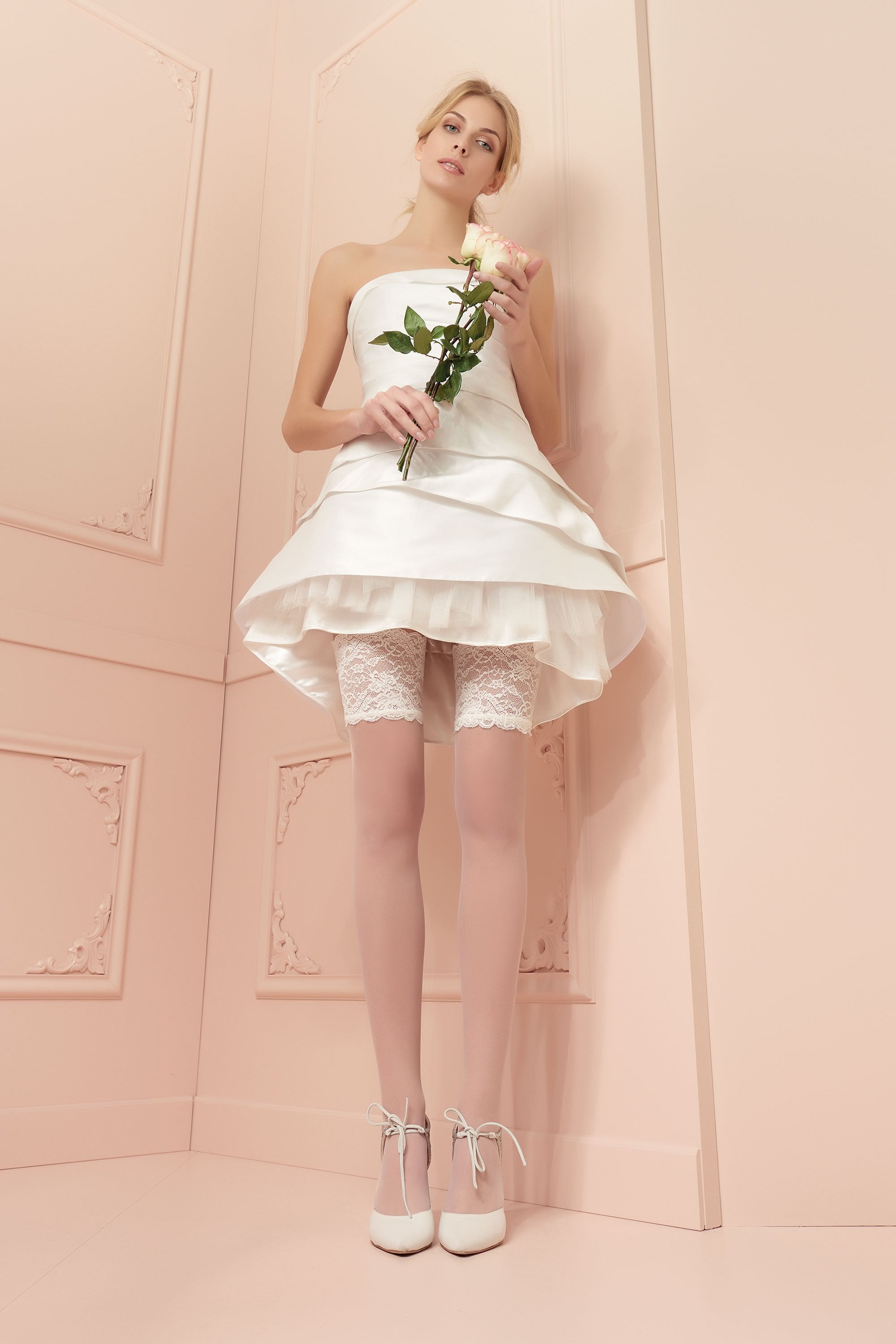 Woman in a white dress and white stay ups holding a rose against a pink wall