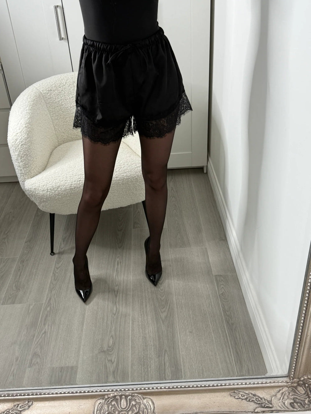 Black lace-trimmed shorts and sheer black tights worn by a model standing in a room with a white armchair and light wood floor, viewed from behind.