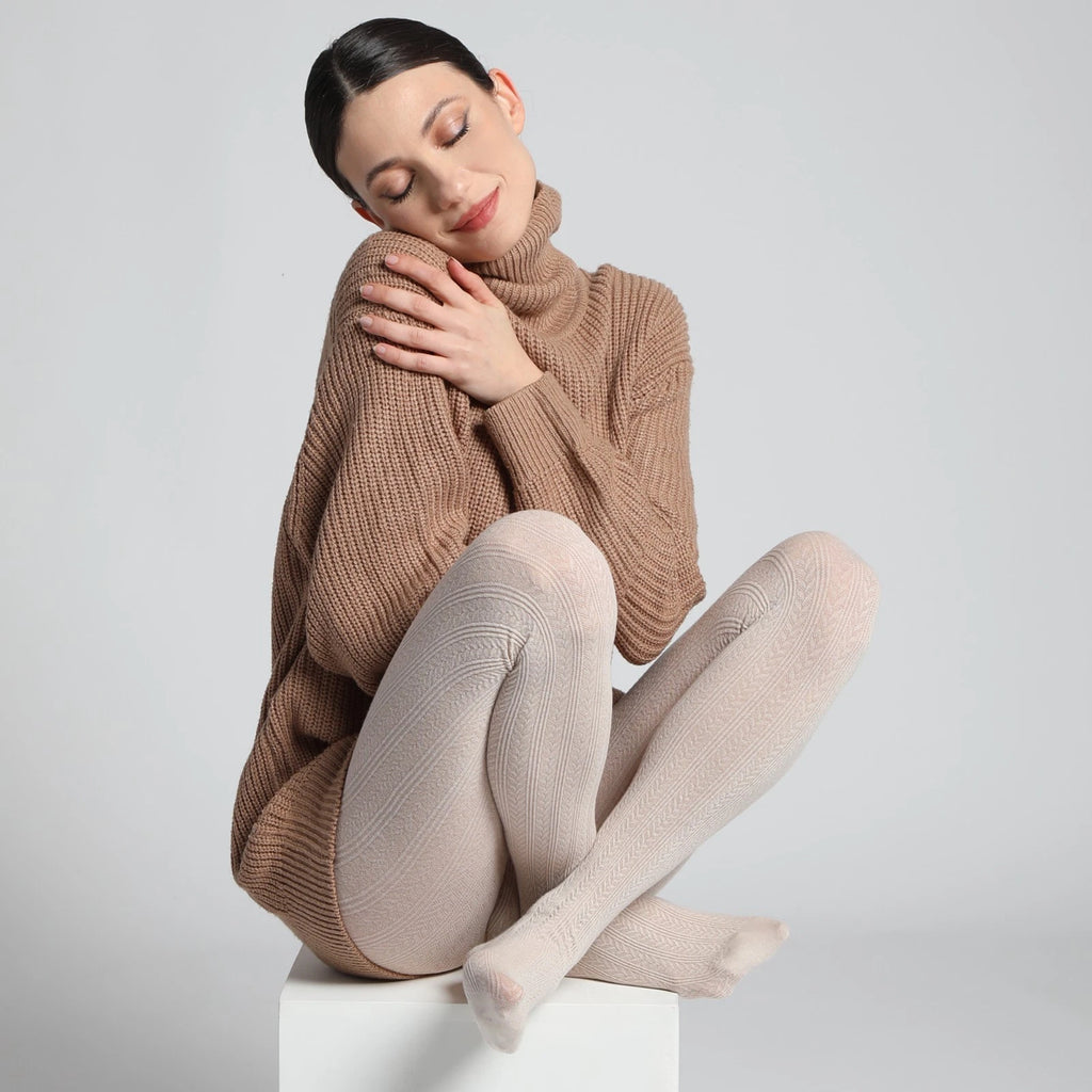 A woman sits cross-legged on a white pedestal against a light gray studio background, wearing an oversized ribbed brown turtleneck sweater and pale blush-beige tights.