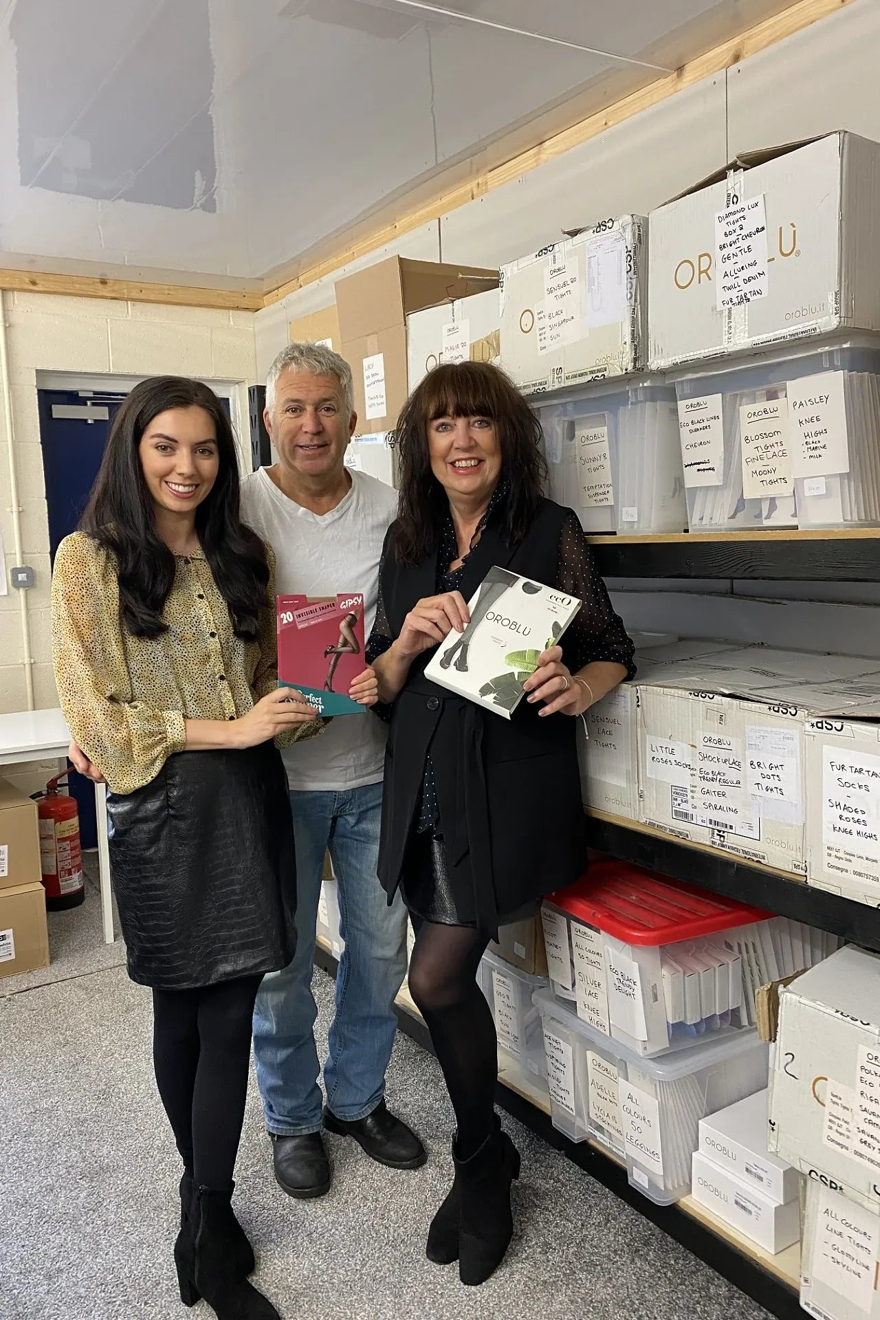 Three people standing in a tights warehouse. The two women are holding up packets of tights.